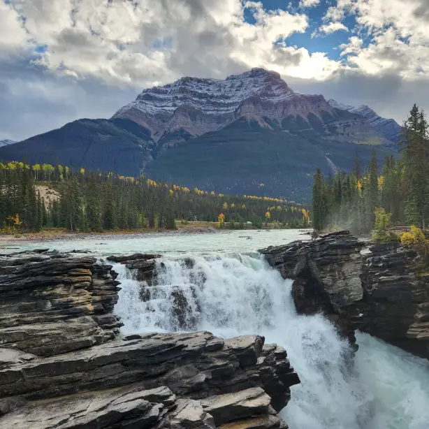 A river flows into a cascade with trees and mountains in the background in Banff National Park, Canada