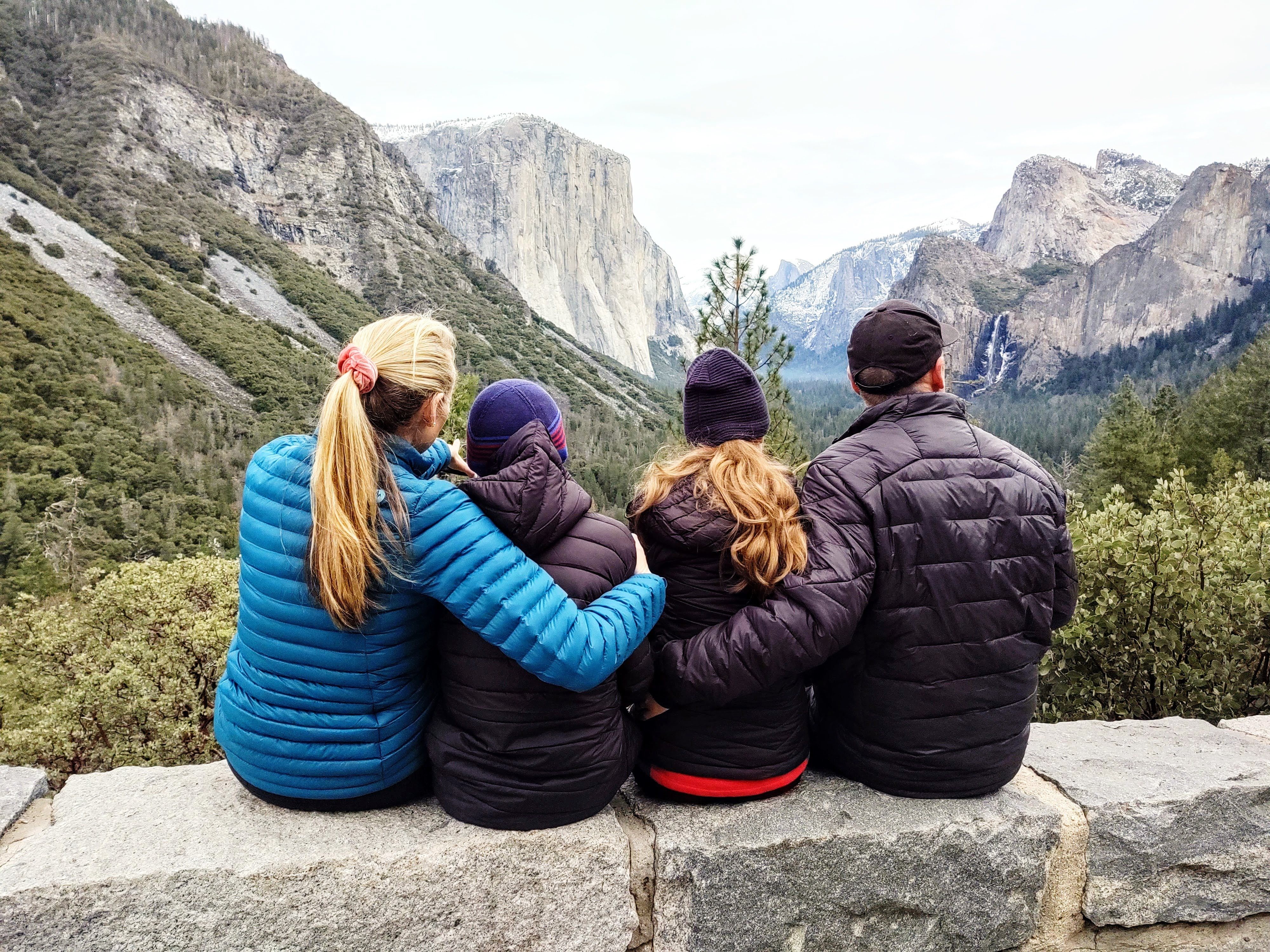 Eine vierköpfige Familie sitzt gemeinsam auf einer Mauer im Yosemite-Nationalpark und genießt den Blick auf hoch aufragende Granitfelsen und Wasserfälle