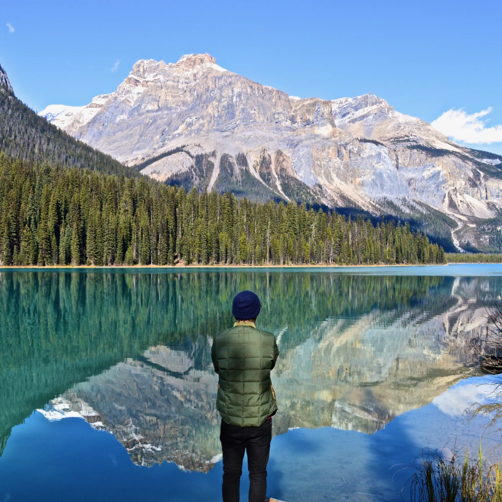 Eine Person steht an einem türkisfarbenen See in Kanada und blickt auf das bewaldete Ufer und die hoch aufragenden Gipfel der Rocky Mountains, die sich im stillen Wasser spiegeln.