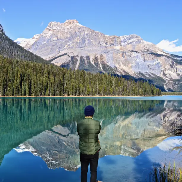 Eine Person steht an einem türkisfarbenen See in Kanada und blickt auf das bewaldete Ufer und die hoch aufragenden Gipfel der Rocky Mountains, die sich im stillen Wasser spiegeln.