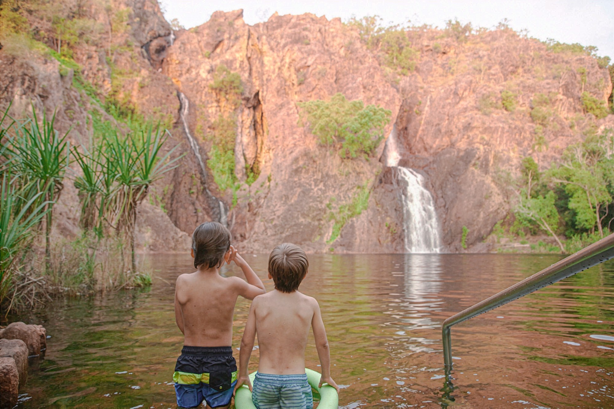 Twee kinderen die in een natuurlijk rotsbad staan en uitkijken naar een waterval die langs rode zandstenen rotswanden naar beneden stort bij Wangi Falls, in het Litchfield National Park, Northern Territory, Australië