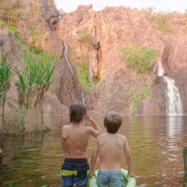 Zwei Kinder waten in einem natürlichen Felsenbecken und blicken auf einen Wasserfall, der an den roten Sandsteinfelsen der Wangi Falls im Litchfield-Nationalpark im Northern Territory, Australien, herabstürzt