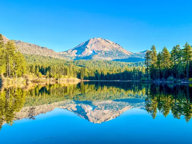 Ein ruhiger See in Kalifornien, in dem sich die umliegenden Kiefernwälder und der markante Vulkangipfel, der sich in der Ferne erhebt, unter einem klaren blauen Himmel spiegeln.
