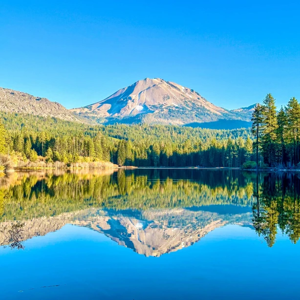 Ein ruhiger See in Kalifornien, in dem sich die umliegenden Kiefernwälder und der markante Vulkangipfel, der sich in der Ferne erhebt, unter einem klaren blauen Himmel spiegeln.