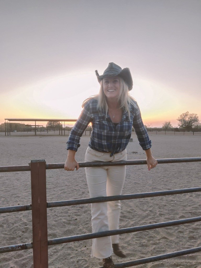 Vrouw met een cowboyhoed, die op een hek leunt.