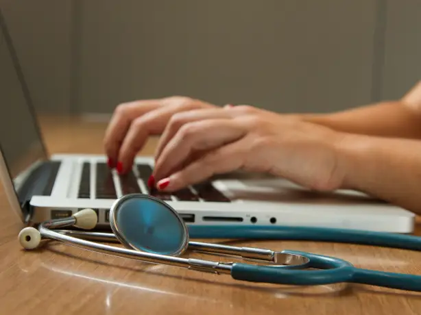 Hände mit rotem Nagellack tippen auf Laptop-Tastatur, daneben liegt ein Stethoskop