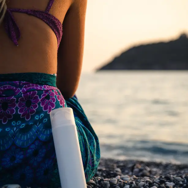Frau von hinten, die auf Kieselstrand und Gewässer sitzt mit einer Sprayflasche an ihr gelehnt