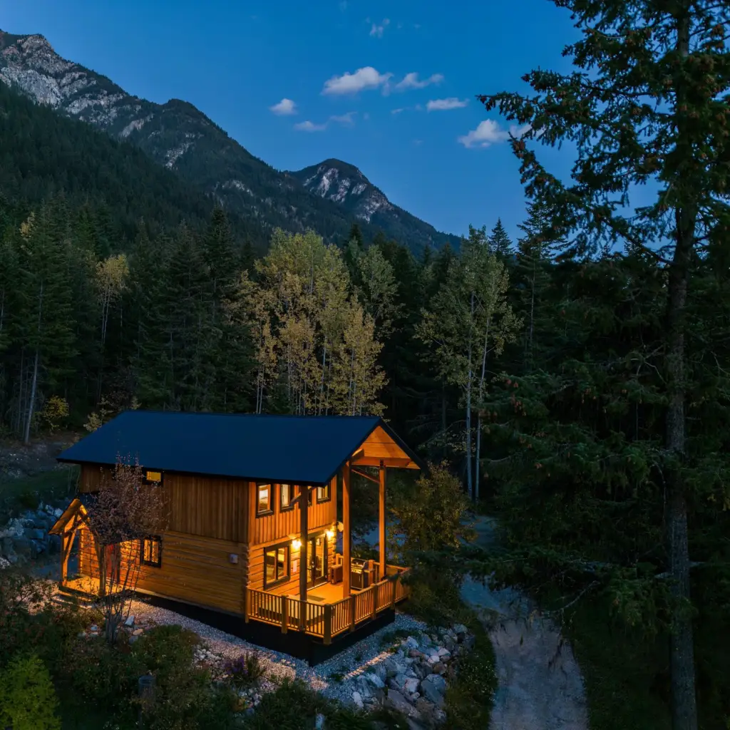 Luchtfoto van een warm verlichte houten hut met overdekt terras, verscholen tussen naaldbomen en espen in de schemering, met ruige bergtoppen op de achtergrond in Golden, Brits-Columbia, Canada.