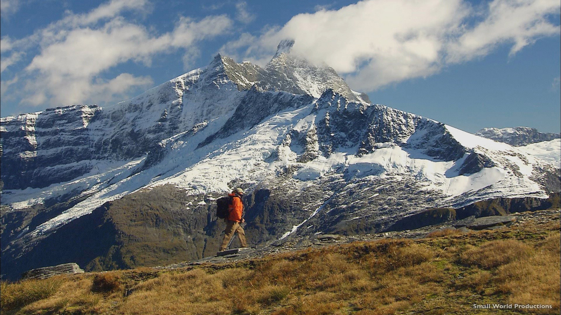 Persoon in een oranje jas loopt door de bergen van Mt. Aspiring National Park met besneeuwde bergtoppen op de achtergrond