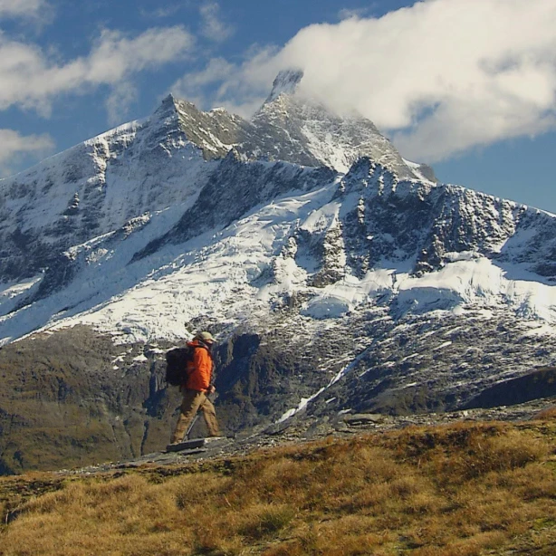 Persoon in een oranje jas loopt door de bergen van Mt. Aspiring National Park met besneeuwde bergtoppen op de achtergrond