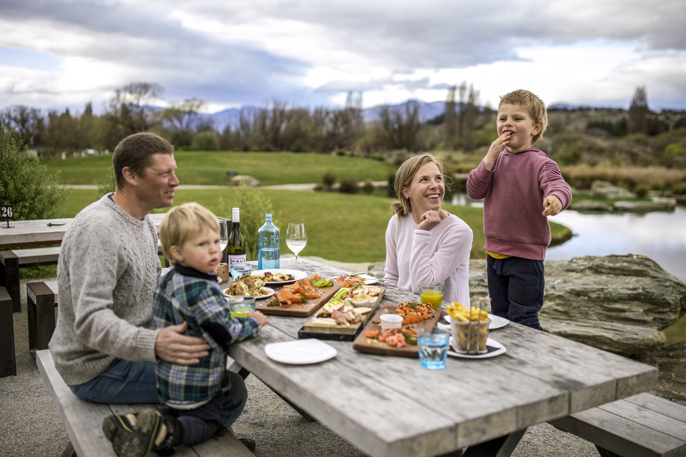 A family sitting down at a picnic table to eat food laid out on wooden boards in Wanaka, New Zealand