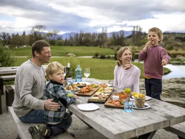 A family sitting down at a picnic table to eat food laid out on wooden boards in Wanaka, New Zealand