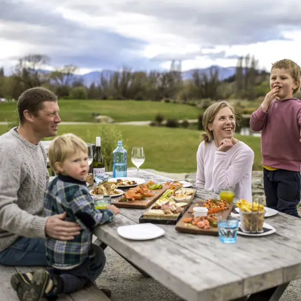 A family sitting down at a picnic table to eat food laid out on wooden boards in Wanaka, New Zealand