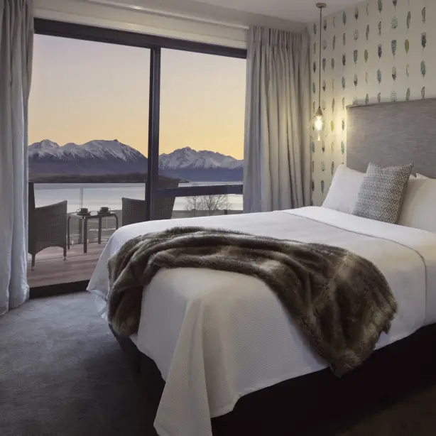 Stylish bed and breakfast room in New Zealand with a king bed, feather-print wallpaper and fur throw, with a private balcony overlooking a lake and snow-capped mountains at sunset
