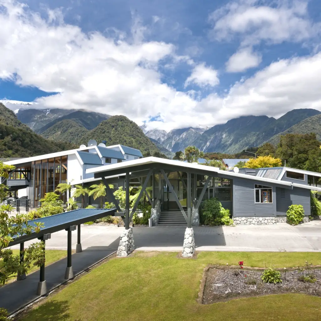 Modernes Boutique-Hotel in Neuseeland, umgeben von üppigen Gärten und einheimischen Farnen, mit der dramatischen Kulisse der schneebedeckten Südlichen Alpen unter einem teilweise bewölkten Himmel