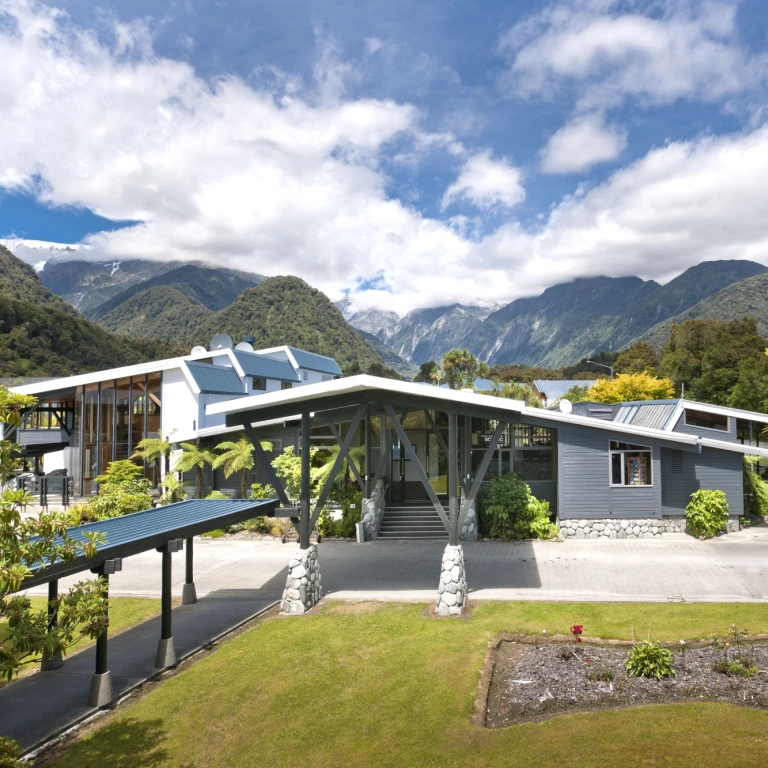 Modern boutique hotel in New Zealand surrounded by lush gardens and native ferns, with a dramatic backdrop of snow-capped Southern Alps mountains under a partly cloudy sky