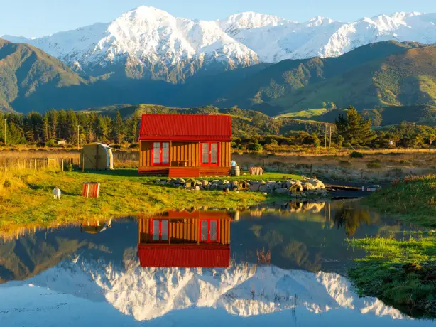 Charmantes Holzcottage mit rotem Dach, das sich in einem ruhigen Teich bei Kaikoura, Neuseeland, spiegelt, mit schneebedeckten Berggipfeln und grünen Hügeln im Hintergrund im goldenen Abendlicht