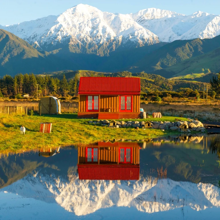 Charmantes Holzcottage mit rotem Dach, das sich in einem ruhigen Teich bei Kaikoura, Neuseeland, spiegelt, mit schneebedeckten Berggipfeln und grünen Hügeln im Hintergrund im goldenen Abendlicht