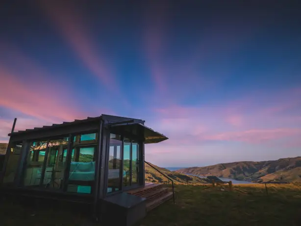 Zeitgenössisches Cottage mit Glaswänden auf einem Hügel in Neuseeland mit Panoramablick über eine Küstenbucht und sanfte Hügel unter einem dramatischen rosa-blauen Abendhimmel