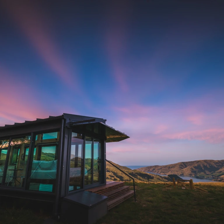 Contemporary glass-walled cottage perched on a hillside in New Zealand with panoramic views over a coastal inlet and rolling hills under a dramatic pink and blue sunset sky