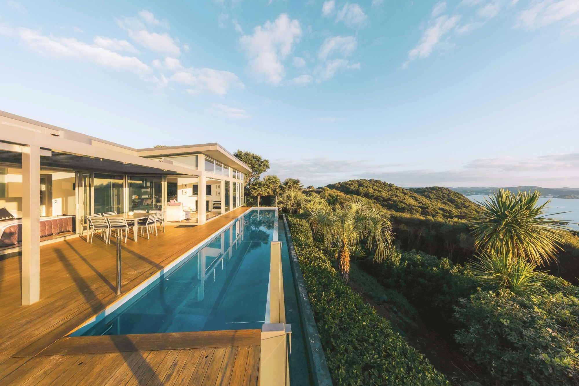 Modern glass house with infinity pool overlooking green hills and water at sunset.