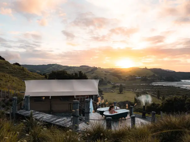 Frau in einer freistehenden Badewanne auf einer rustikalen Terrasse neben einem Glamping-Zelt in Neuseeland, mit weitem Blick über sanfte grüne Hügel und eine Küstenbucht bei Sonnenuntergang