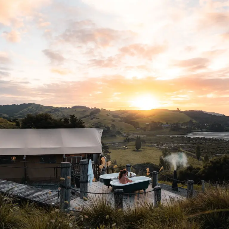 Frau in einer freistehenden Badewanne auf einer rustikalen Terrasse neben einem Glamping-Zelt in Neuseeland, mit weitem Blick über sanfte grüne Hügel und eine Küstenbucht bei Sonnenuntergang