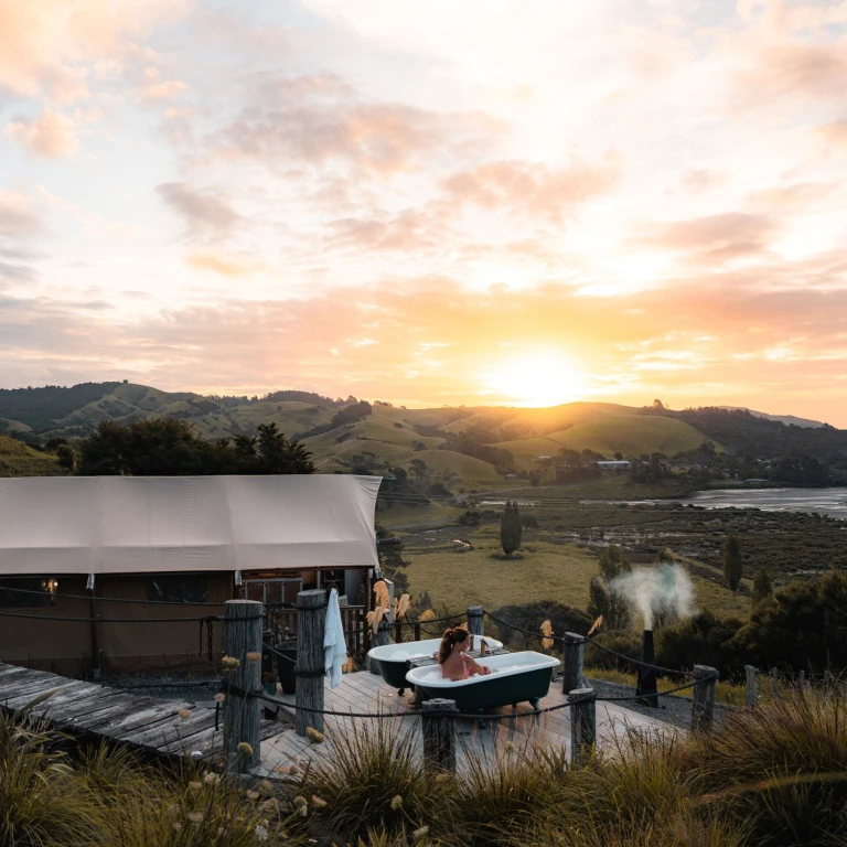 Frau in einer freistehenden Badewanne auf einer rustikalen Terrasse neben einem Glamping-Zelt in Neuseeland, mit weitem Blick über sanfte grüne Hügel und eine Küstenbucht bei Sonnenuntergang