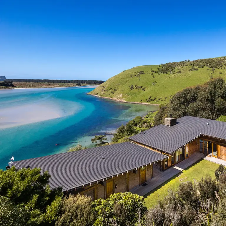 Abgelegenes Holzferienhaus über einer türkisfarbenen Küstenbucht in Neuseeland, umgeben von einheimischem Buschland und sanften grünen Hügeln unter einem klaren blauen Himmel