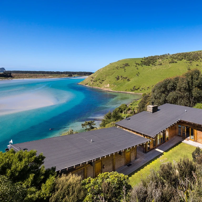 Secluded timber holiday house perched above a turquoise coastal inlet in New Zealand, surrounded by native bush and rolling green hills under a clear blue sky