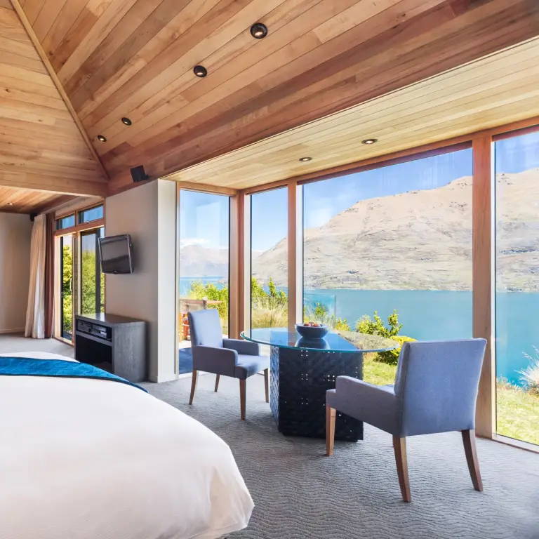 Ruime lodgesuite in Nieuw-Zeeland met een gewelfd houten plafond, kingsize bed, open haard en kamerhoge ramen met uitzicht op een turquoise meer en berglandschap.
