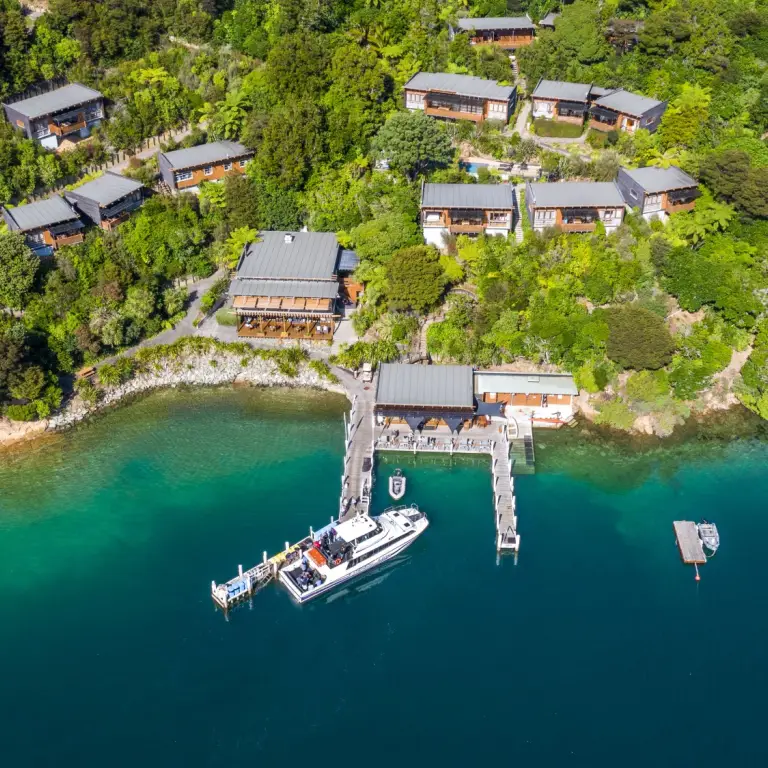 Luftaufnahme eines Wasserfront-Lodgeresorts in den Marlborough Sounds, Neuseeland, mit Holzchalets eingebettet in üppiges einheimisches Buschland an einem Hang über einem privaten Steg und türkisfarbenem Meer
