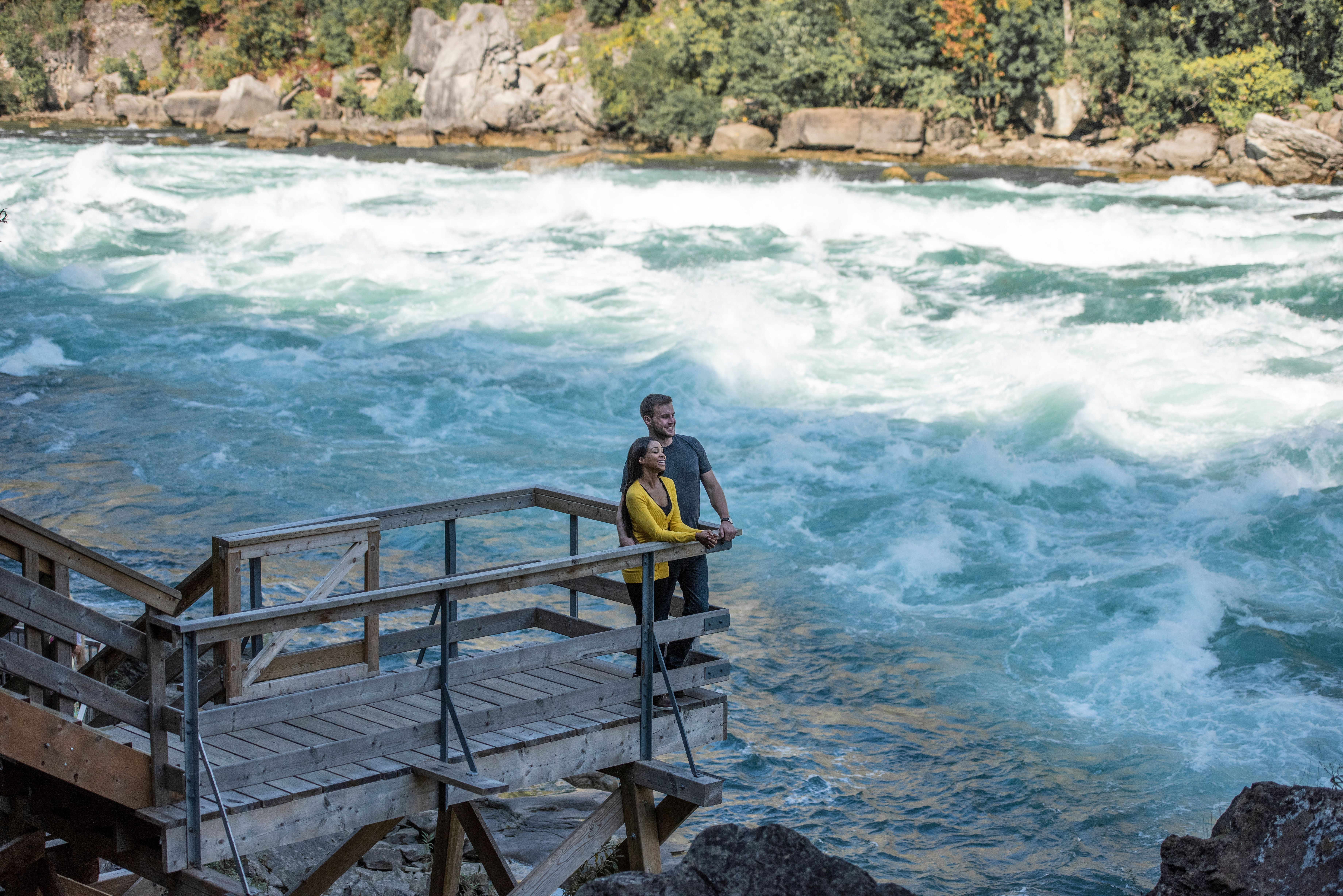 A couple stand on a wooden viewing platform with a fast flowing river running past them at Niagara Falls