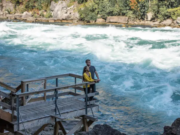 A couple stand on a wooden viewing platform with a fast flowing river running past them at Niagara Falls