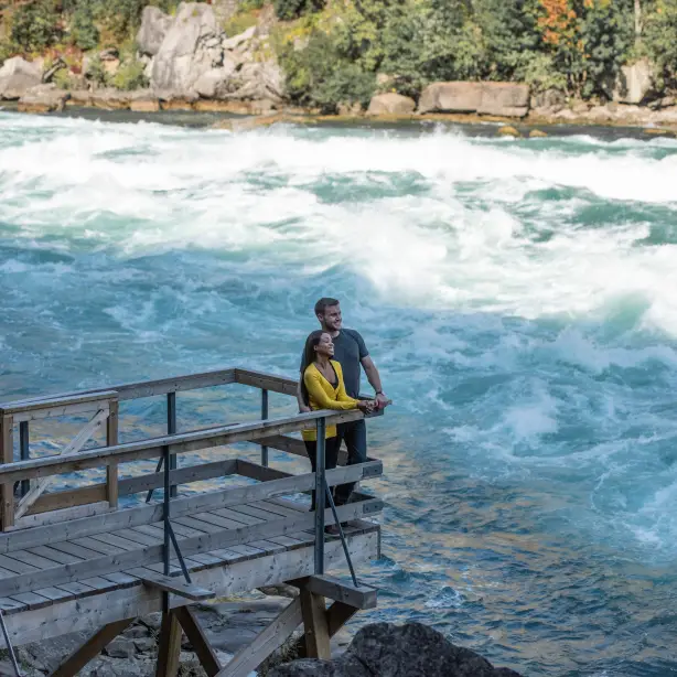 A couple stand on a wooden viewing platform with a fast flowing river running past them at Niagara Falls