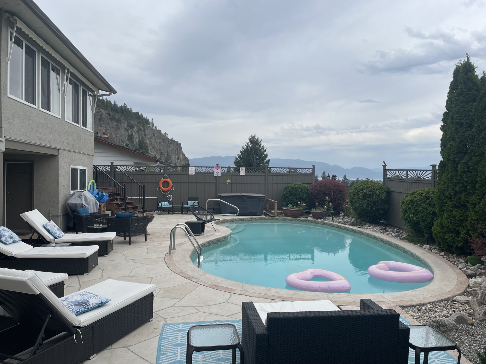 Een privé buitenzwembad met roze opblaasbanden en ligbedden op een betegeld terras, met een jacuzzi, tuinstoelen en uitzicht op het Okanagan Lake en de omliggende bergen achter het hek, bij een villa in de Okanagan Valley, Brits-Columbia, Canada.