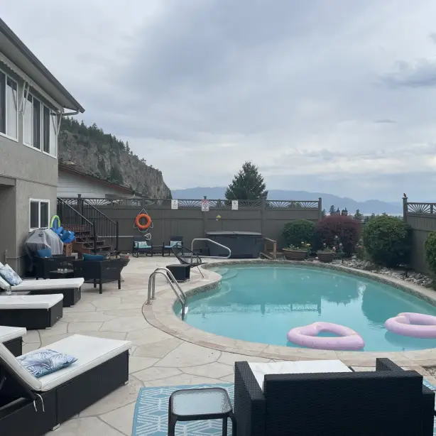 Ein privater Außenpool mit rosa Schwimmringen und Liegestühlen auf einer gepflasterten Terrasse, mit Whirlpool, Gartenmöbeln und Blick auf den Okanagan Lake und die umliegenden Berge hinter dem Zaun, bei einer Villa im Okanagan Valley, British Columbia, Kanada.