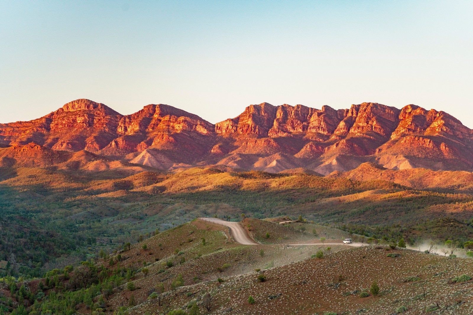 Eine staubige Outback-Piste schlängelt sich durch die Flinders Ranges in Südaustralien, mit zerklüfteten roten Bergketten, die im warmen Licht der Abendsonne leuchten.