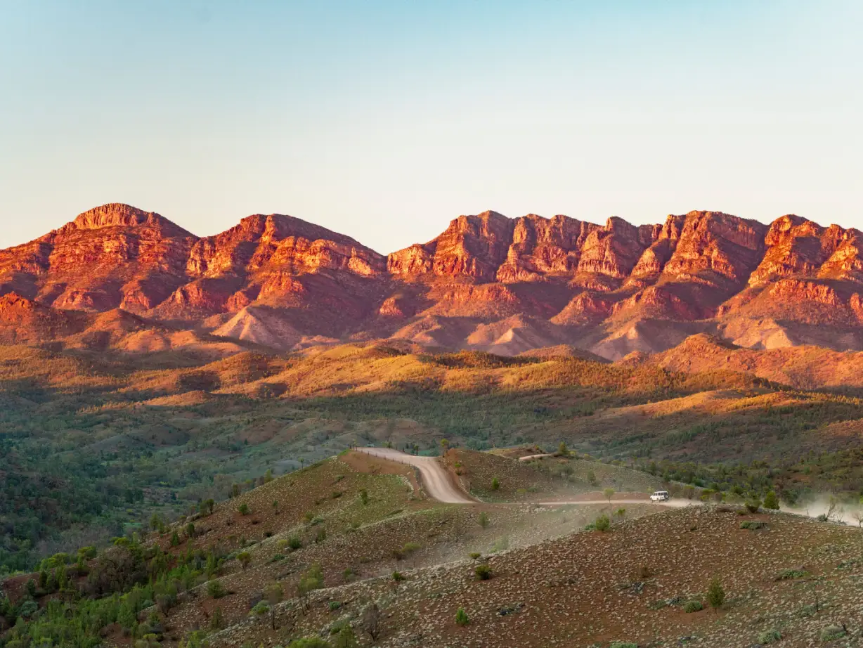 Eine staubige Outback-Piste schlängelt sich durch die Flinders Ranges in Südaustralien, mit zerklüfteten roten Bergketten, die im warmen Licht der Abendsonne leuchten.
