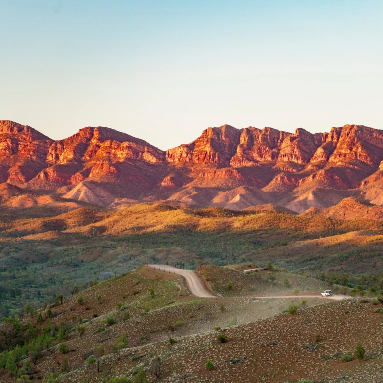 Eine staubige Outback-Piste schlängelt sich durch die Flinders Ranges in Südaustralien, mit zerklüfteten roten Bergketten, die im warmen Licht der Abendsonne leuchten.
