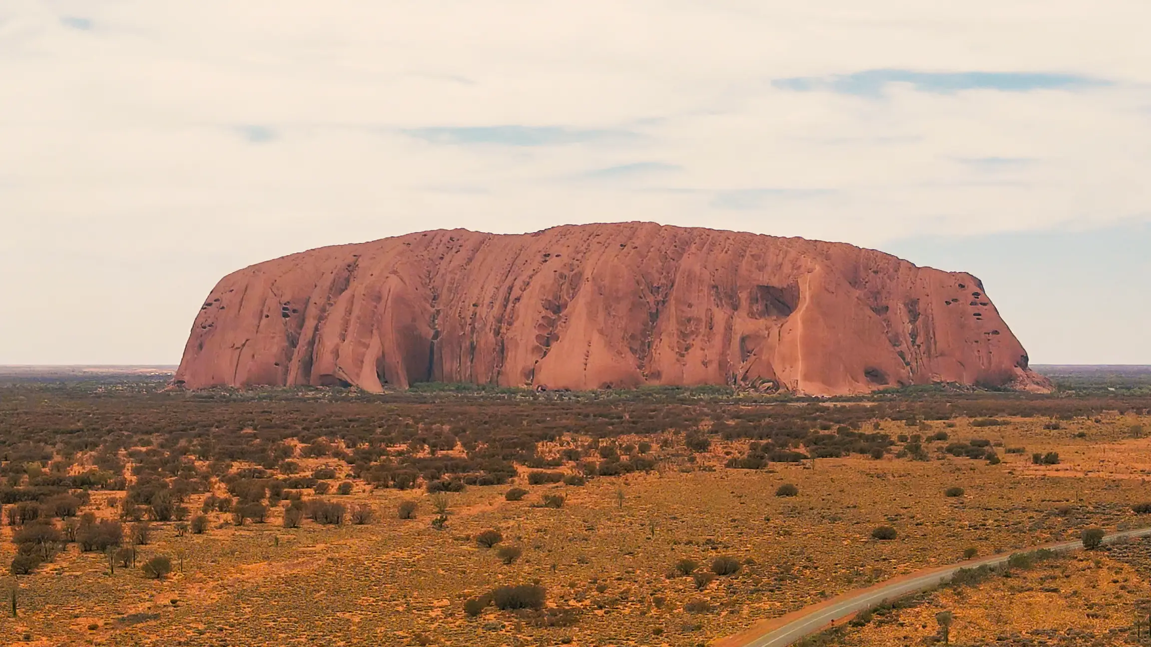 Uluru am Horizont, ein Auto fährt davor auf einer Straße entlang