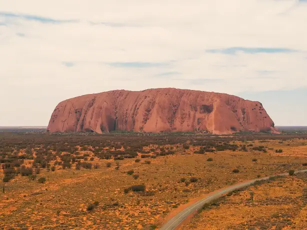 Uluru am Horizont, ein Auto fährt davor auf einer Straße entlang