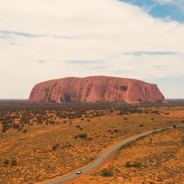 Uluru am Horizont, ein Auto fährt davor auf einer Straße entlang