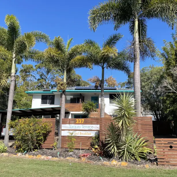 Two-storey bed and breakfast accommodation in Australia, surrounded by tall palm trees and tropical gardens behind a timber fence, under a clear blue sky