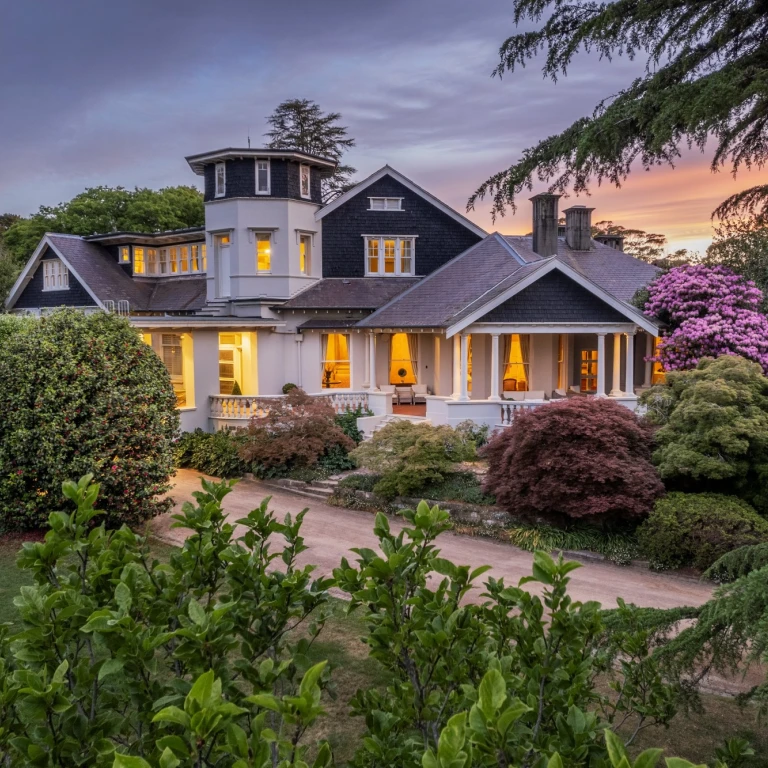 Große historische Bed-and-Breakfast-Unterkunft in Australien, mit einem herrschaftlichen Herrenhaus mit umlaufender Veranda und Turm, umgeben von gepflegten Gärten mit japanischen Ahornbäumen und blühenden Rhododendren bei Sonnenuntergang