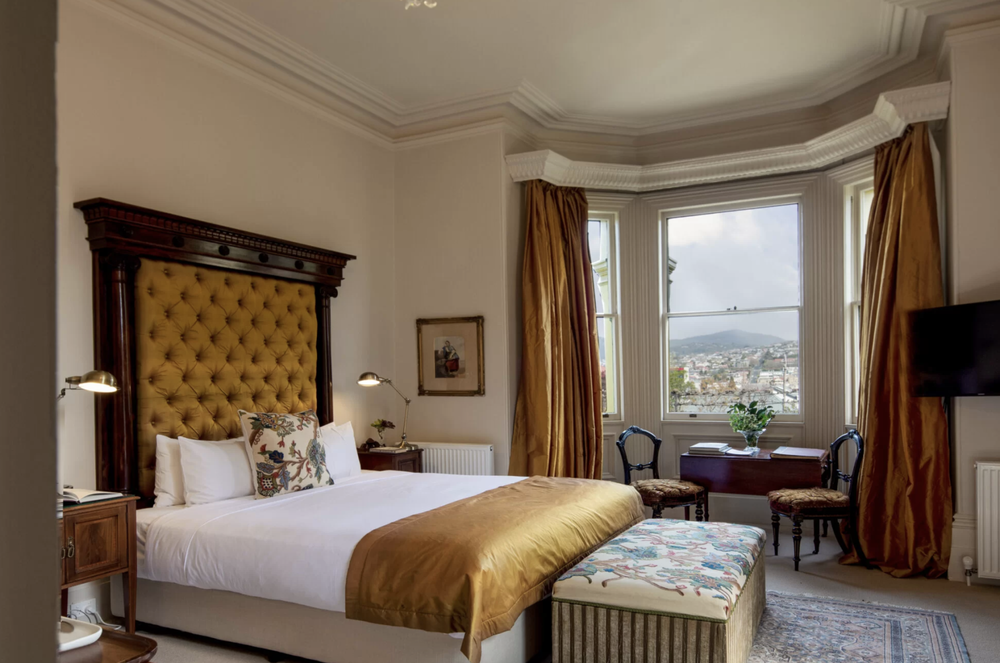 Bedroom with a large upholstered headboard, gold curtains, a window overlooking a hillside town, and antique-style furniture.