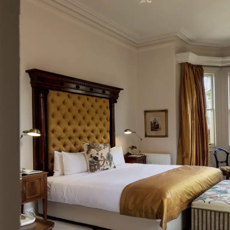 Bedroom with a large upholstered headboard, gold curtains, a window overlooking a hillside town, and antique-style furniture.