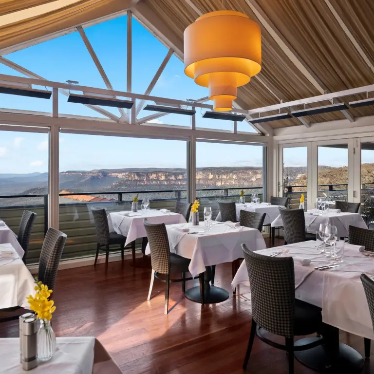 Stijlvol boetiekhotel-restaurant in Australië met witgedekte eettafels en panoramisch uitzicht over de Blue Mountains door kamerhoge ramen onder een gewelfd houten plafond en bergen op de achtergrond.