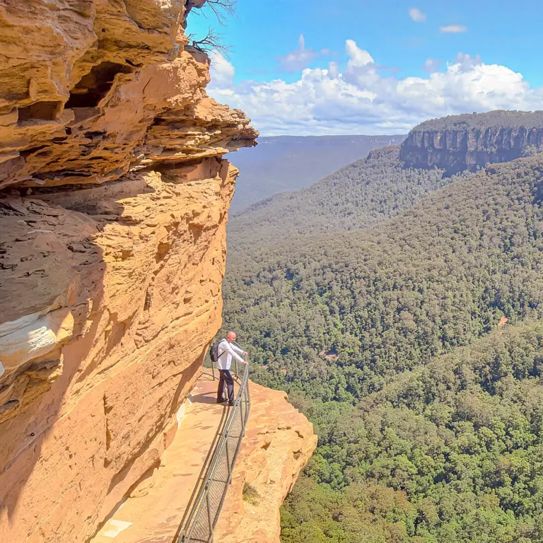 Hiker standing on a cliffside walkway overlooking the vast forested valleys and sandstone escarpments of the Blue Mountains, New South Wales, Australia