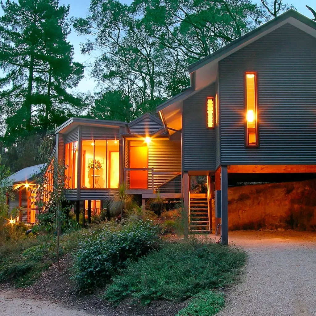 Zeitgenössisches Cottage auf Stelzen mit Wellblechverkleidung und warm leuchtendem Innenraum, eingebettet zwischen hohen Bäumen im australischen Busch in der Abenddämmerung